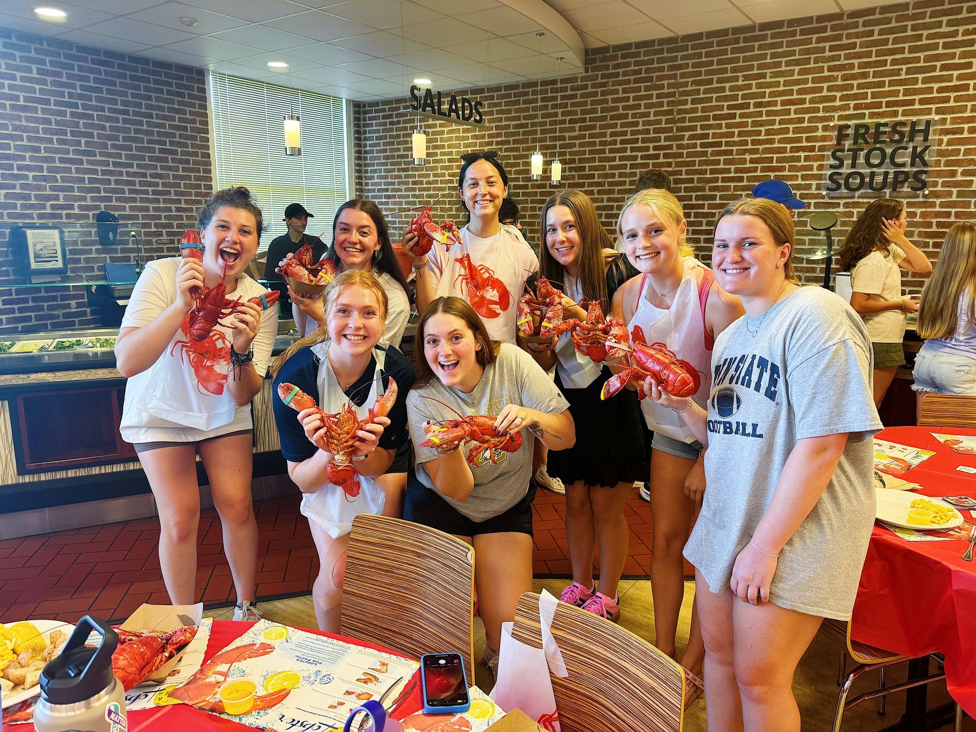 Group of smiling young women holding cooked lobsters at a buffet.