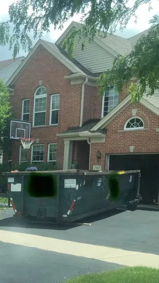 A large dumpster sits in the driveway of a two-story brick home, with a basketball hoop mounted on the house exterior.