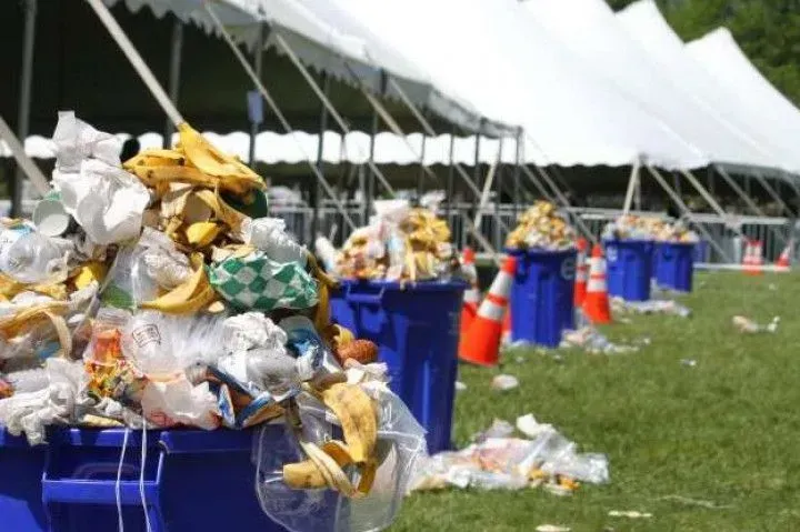 Blue trash bins overflowing with food waste and litter sit in a grassy area lined with white event tents.