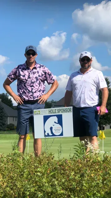 Two individuals in hats and sunglasses pose on a golf course behind a 