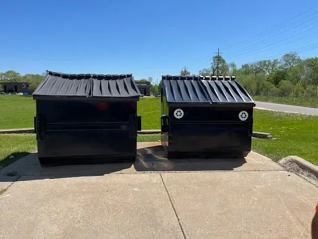 Two black commercial dumpsters with slanted lids sit side-by-side on a concrete pad in an outdoor grassy area.