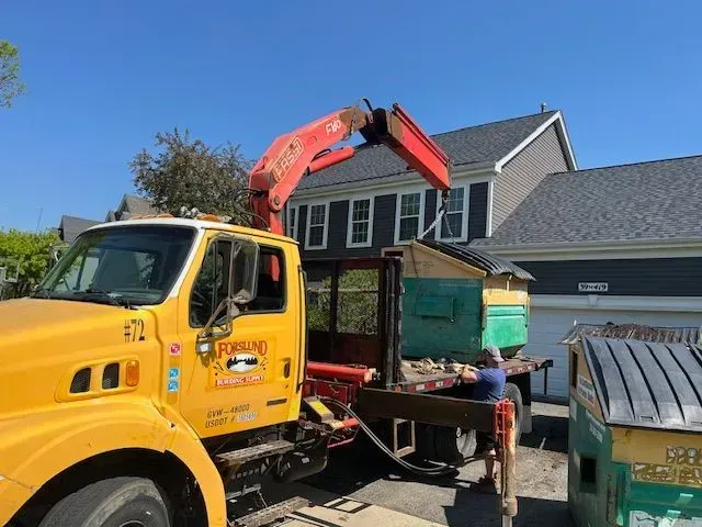 A yellow crane truck with a red boom arm lifts a debris container in front of a house on a sunny day.