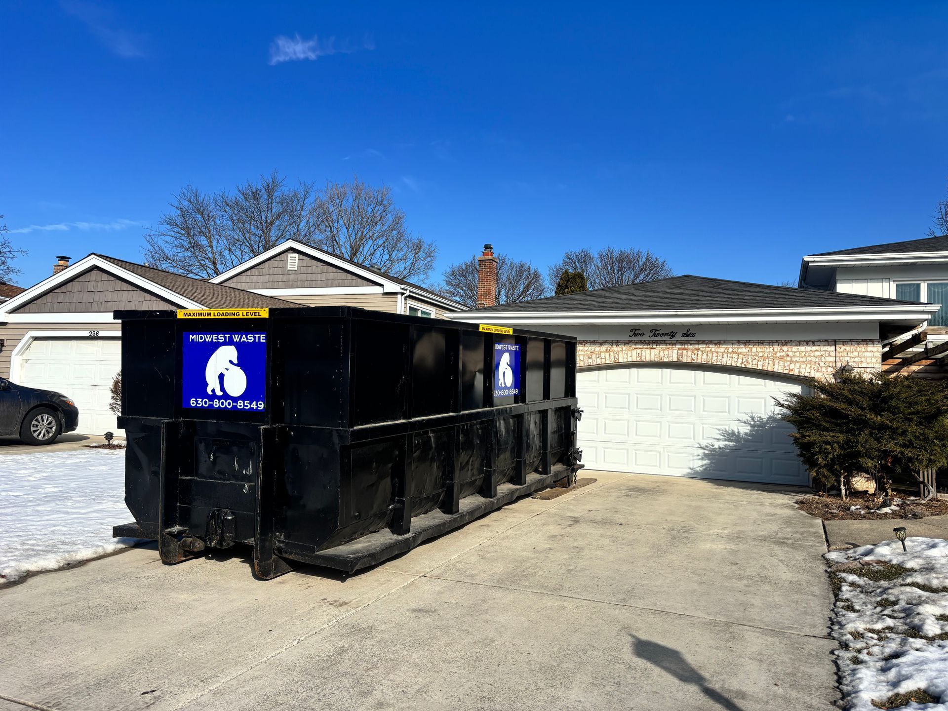 A large black dumpster sits in the concrete driveway of a residential suburban house.
