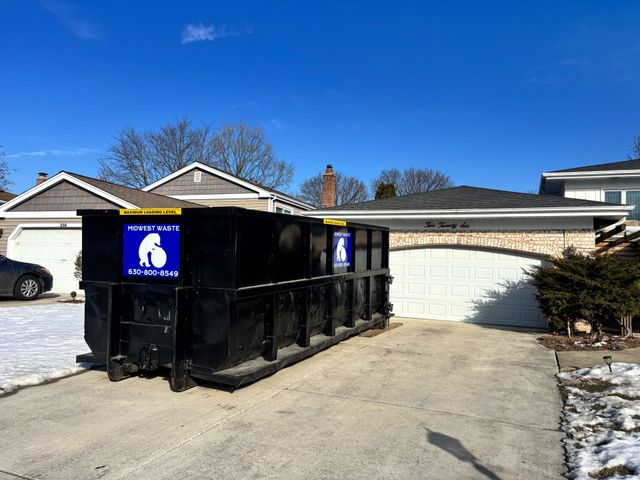 A large black dumpster sits in the concrete driveway of a residential suburban house.