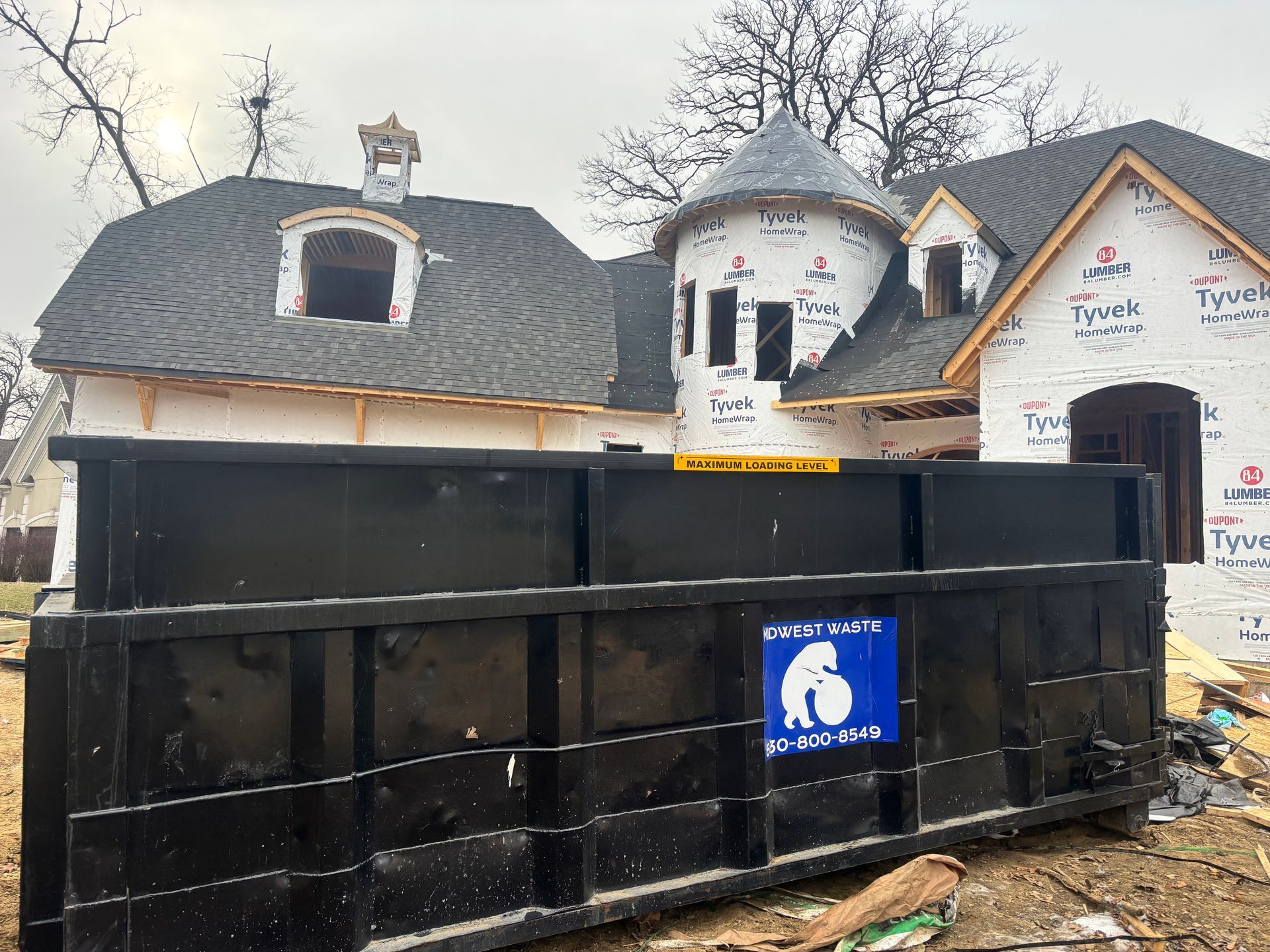 A large black dumpster sits in front of a house under construction, which features a prominent turret and Tyvek wrap.