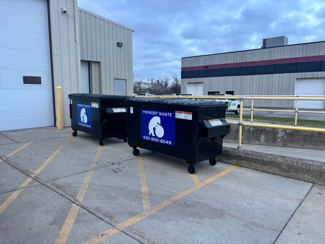 A pile of unsorted paper and plastic waste inside a warehouse, being fed into a large blue industrial baler.