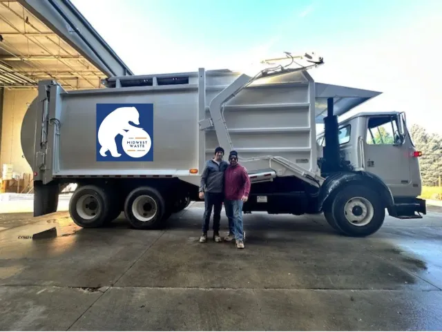 Two people stand beside a large, silver garbage truck featuring a blue logo of a white bear with a sphere.