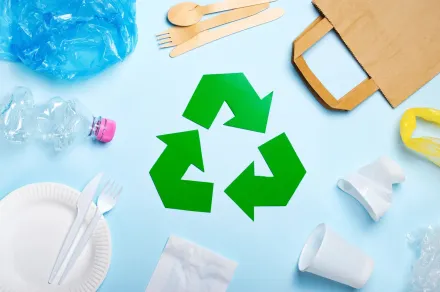 A green recycling symbol is surrounded by plastic , paper , and wooden utensils on a blue surface.