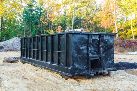 A large black dumpster is sitting in the middle of a dirt field.