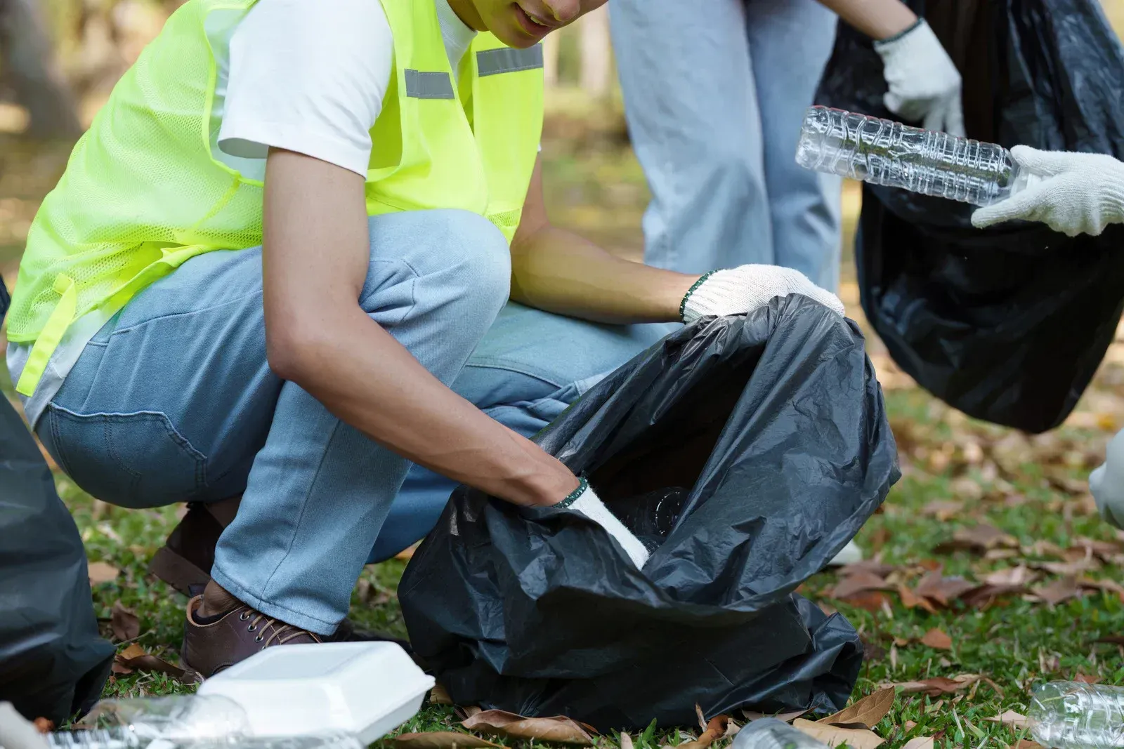 Volunteers wearing reflective vests and gloves collect litter into black plastic bags in a park.