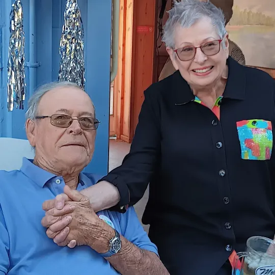 An older couple holding hands. A woman in a black shirt stands beside a man in a blue polo shirt, both smiling at the camera.