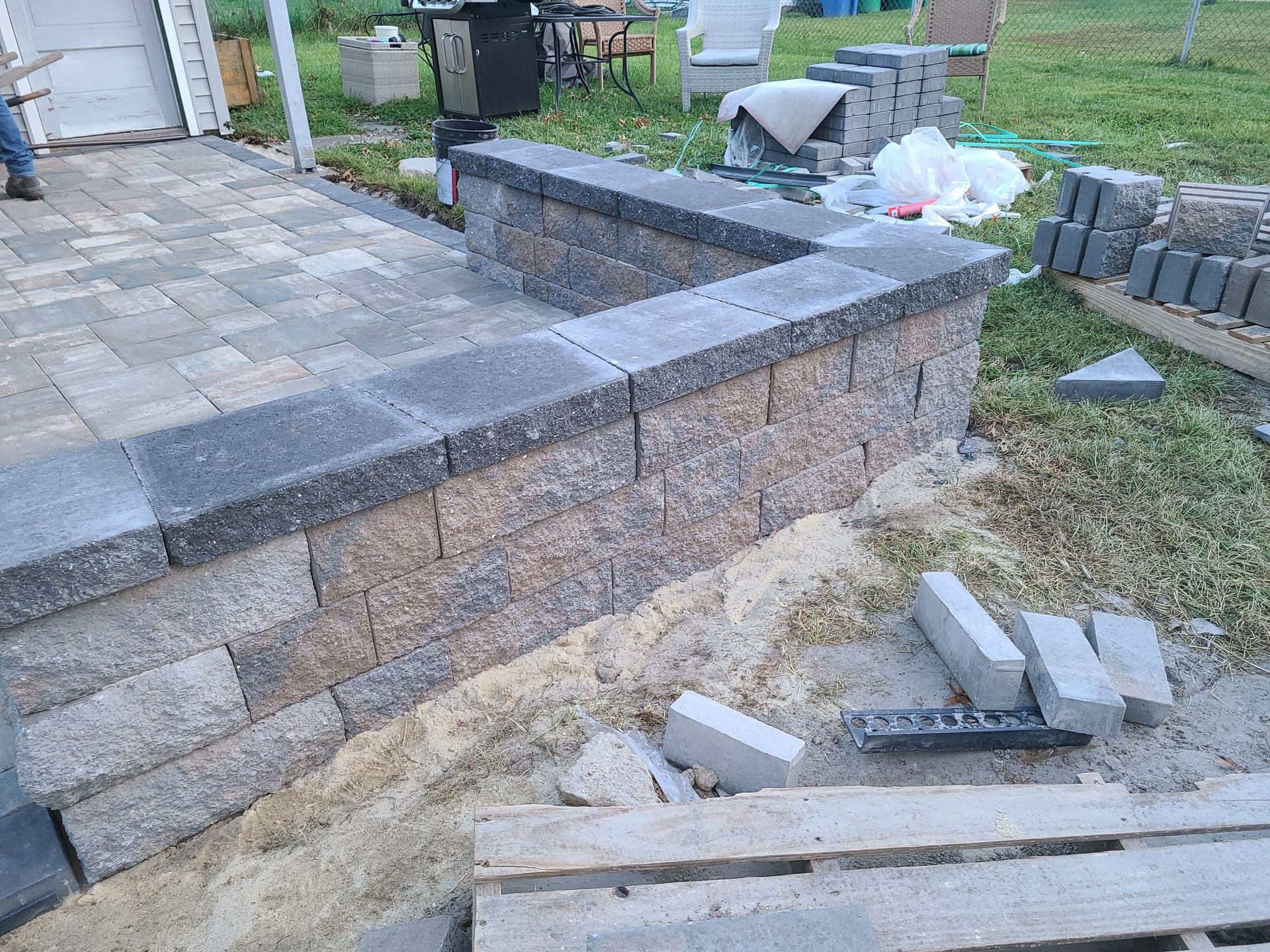 Broken stacked retaining wall blocks beside a stone patio, with loose pavers and debris on the ground.