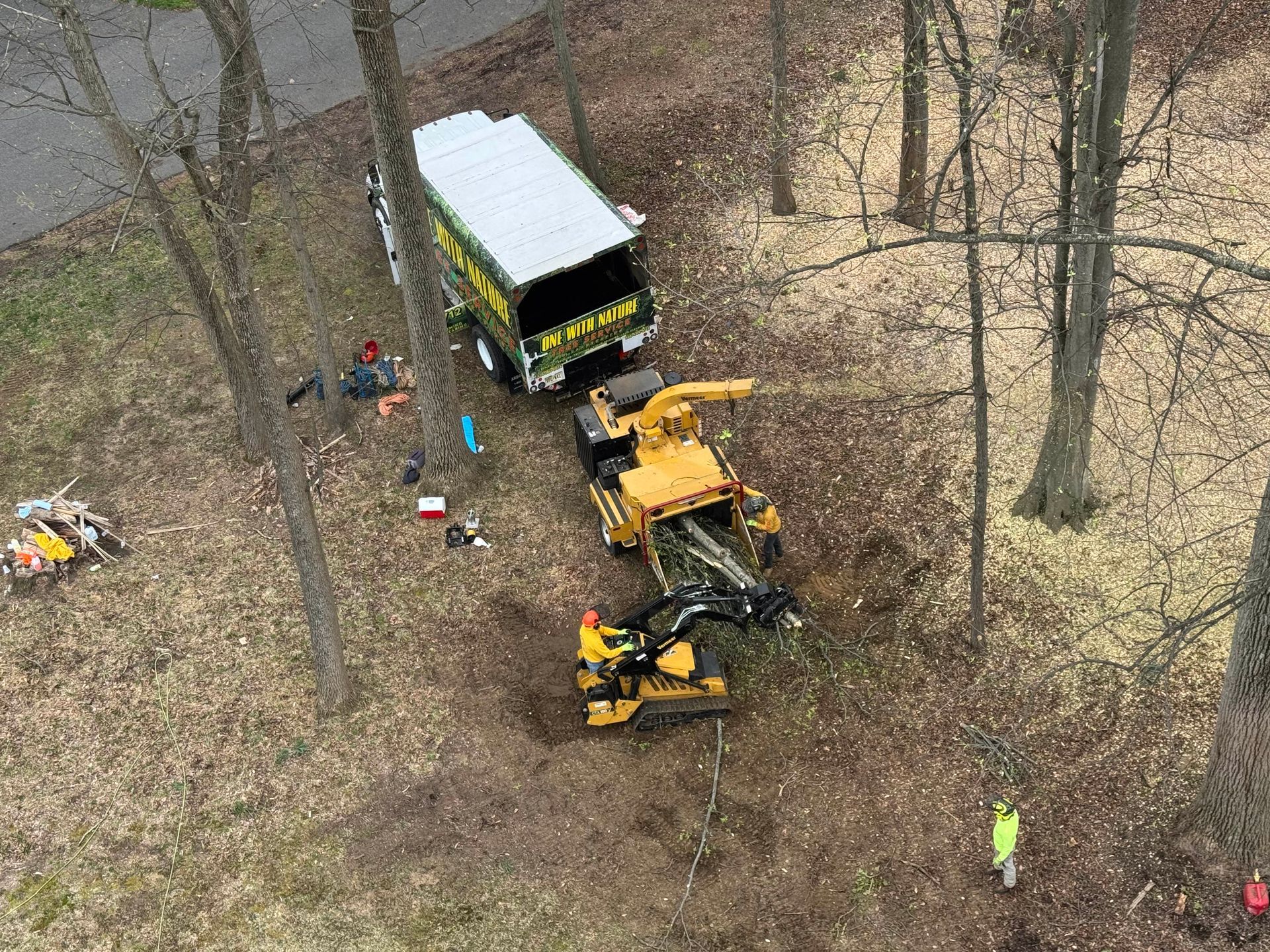 Aerial view of a yellow excavator loading debris into a truck in a wooded area with workers nearby