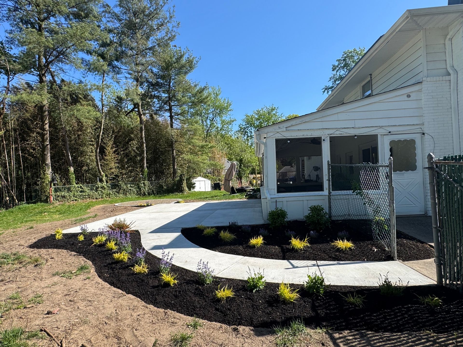White house with screened porch, curved walkway, and new landscaped flower beds beside a wooded yard