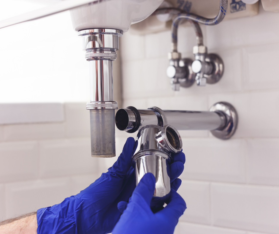 A person wearing blue gloves repairs a chrome sink P-trap pipe under a bathroom vanity.