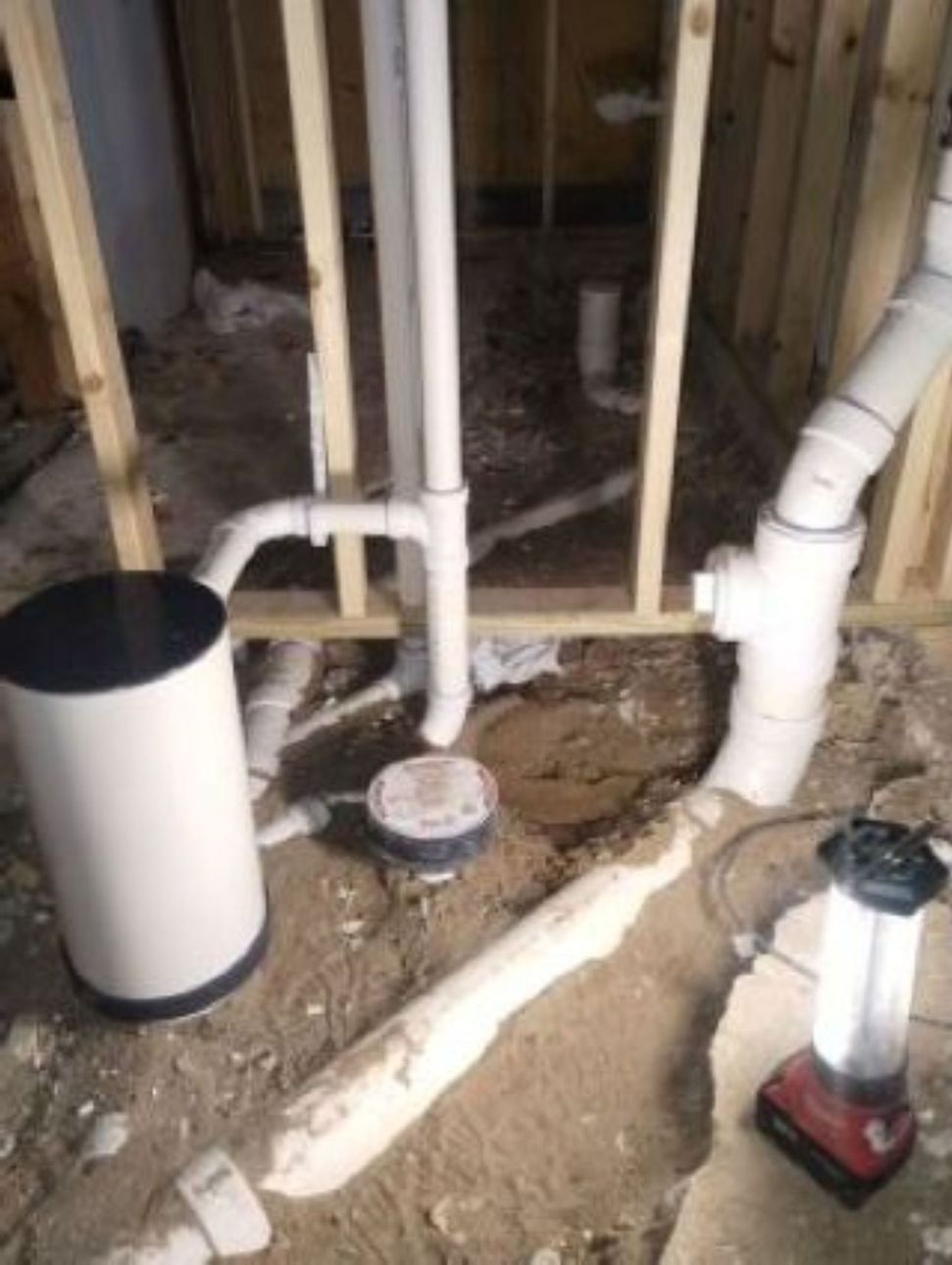 Rough-in plumbing featuring a white sump pump basin, PVC pipes, and a cleanout cap installed in a dirt floor.