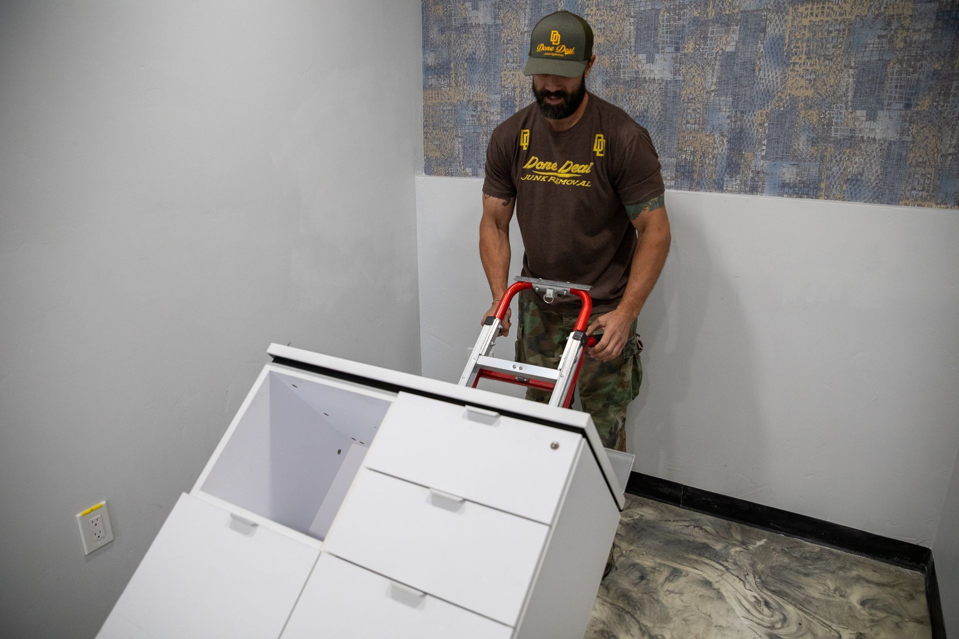 A man with a Done Deal Junk Removal shirt on is rolling a desk out of a room with a dolly.