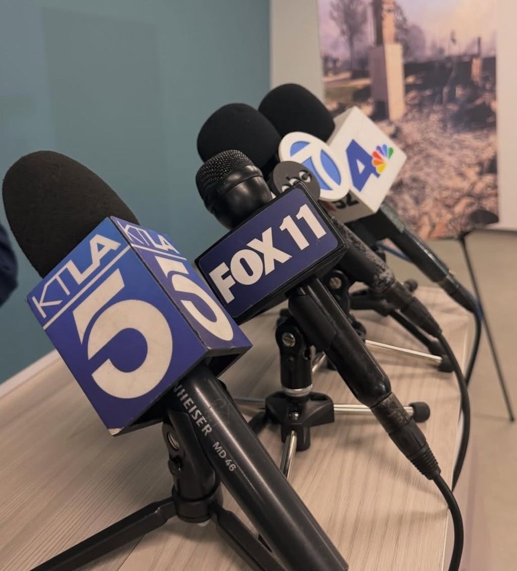 Array of microphones with different station logos, a woman with red hair in the background looking at her phone.