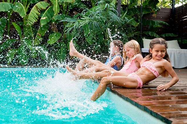 Three young girls splashing in a pool, kicking their feet, smiling. Lush greenery.