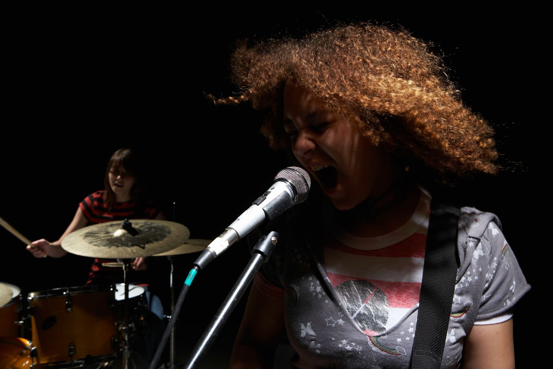 Woman singing into microphone with drummer in background.