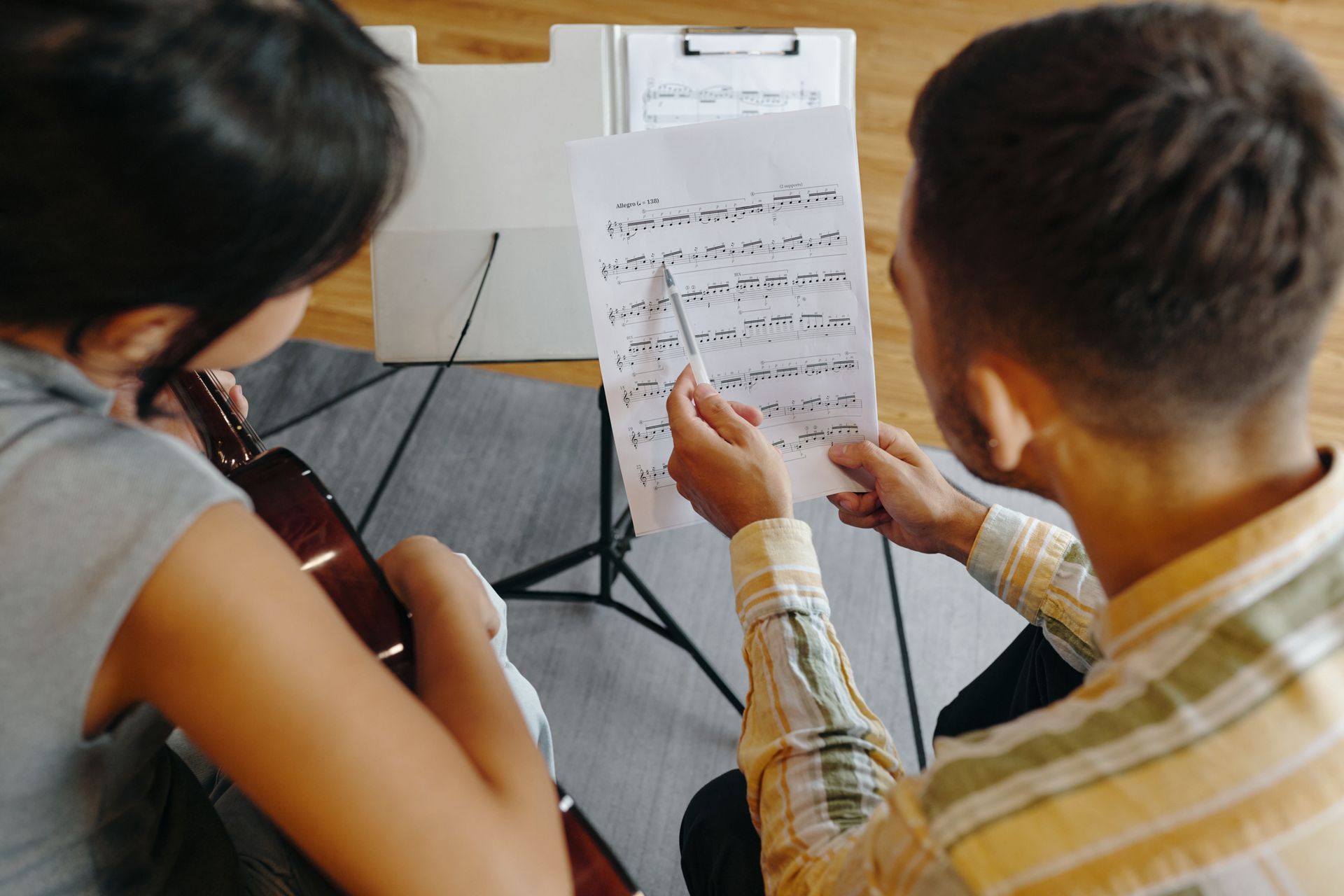 Two people, one with a guitar, studying sheet music together; indoors.