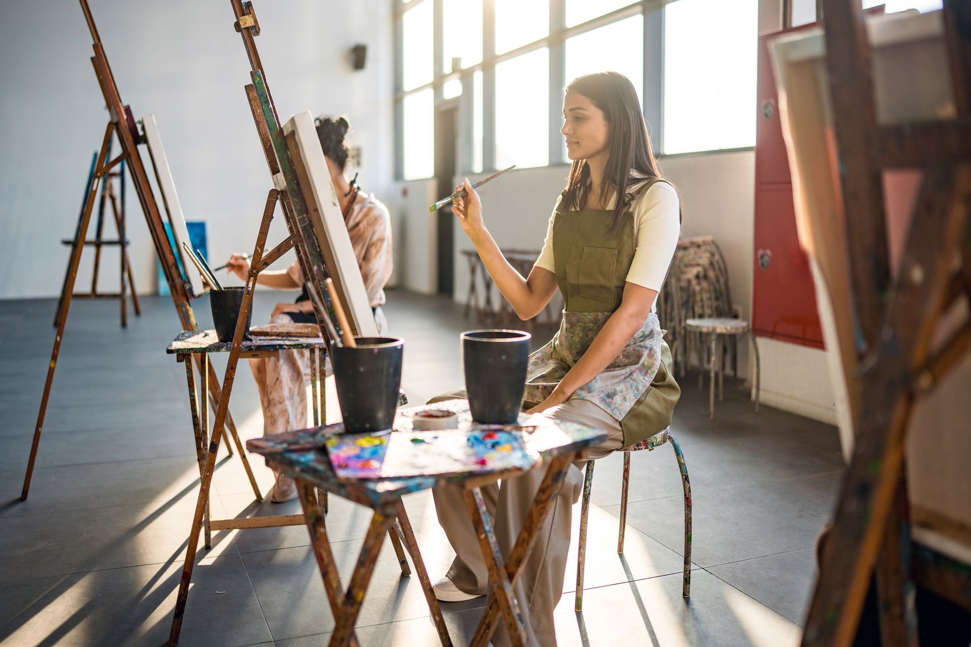 Two women painting in an art studio. One holds a brush, wearing an apron. Sunlight streams in.