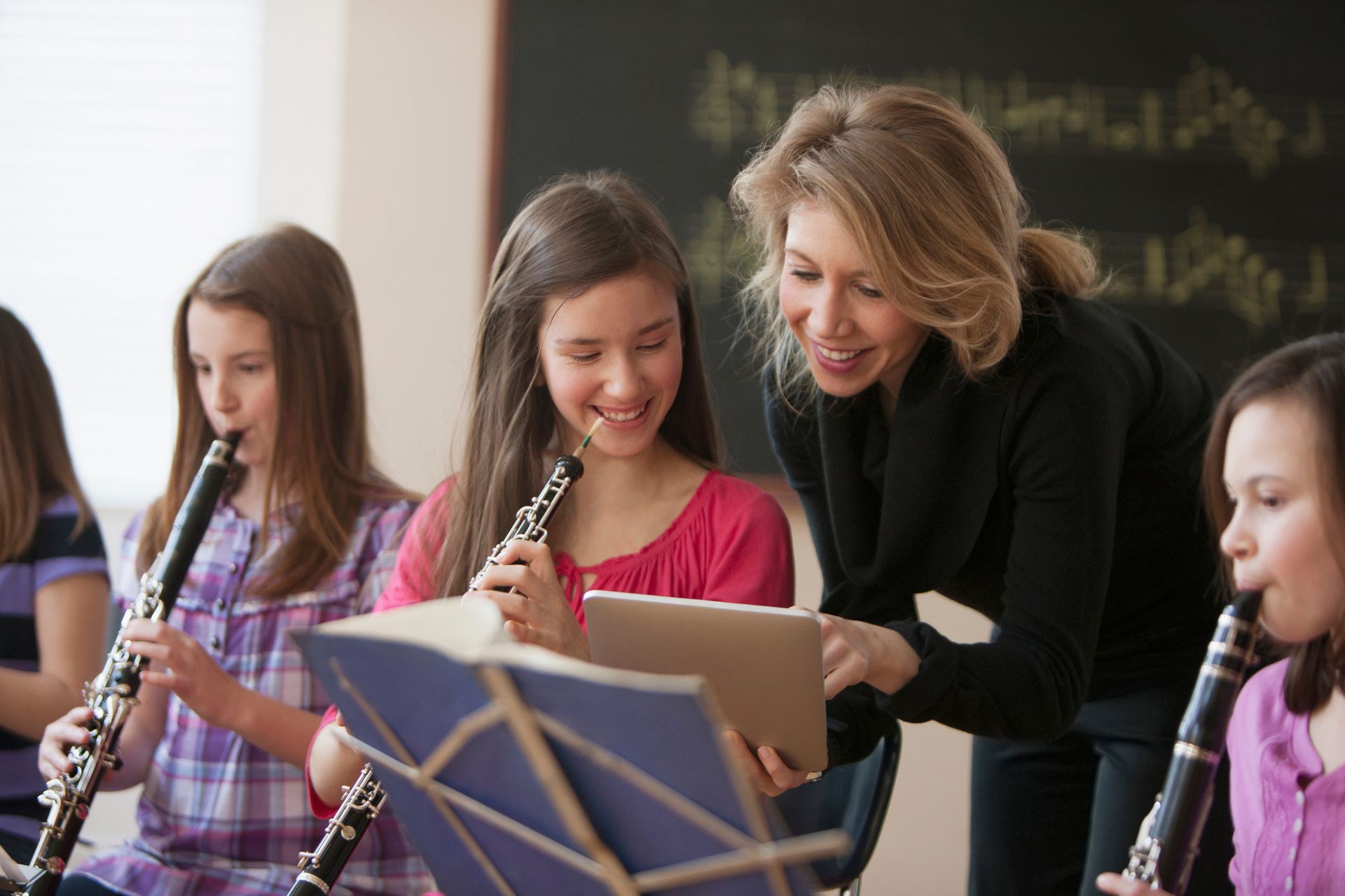 Music teacher helps girl playing the clarinet, smiles, in a classroom with other students.