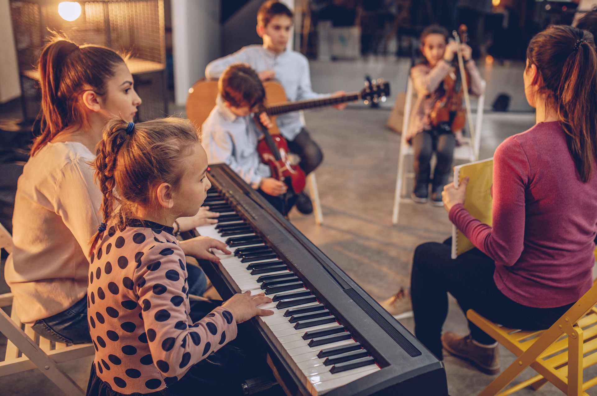 A music class with children playing piano, guitar, violin, and a teacher, indoors.