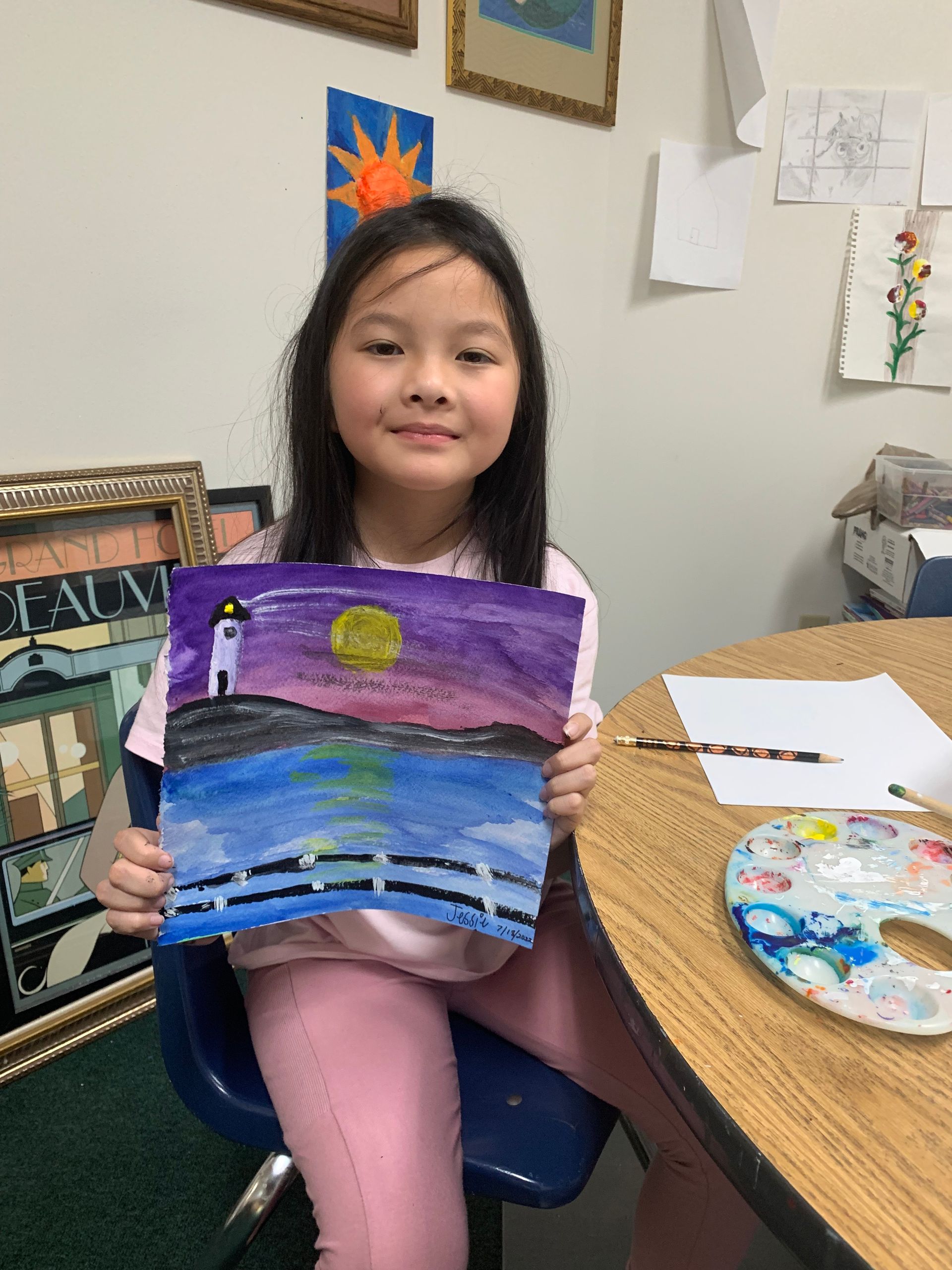 Girl smiling, holding a watercolor painting of a lighthouse at sunset. She's in a classroom.