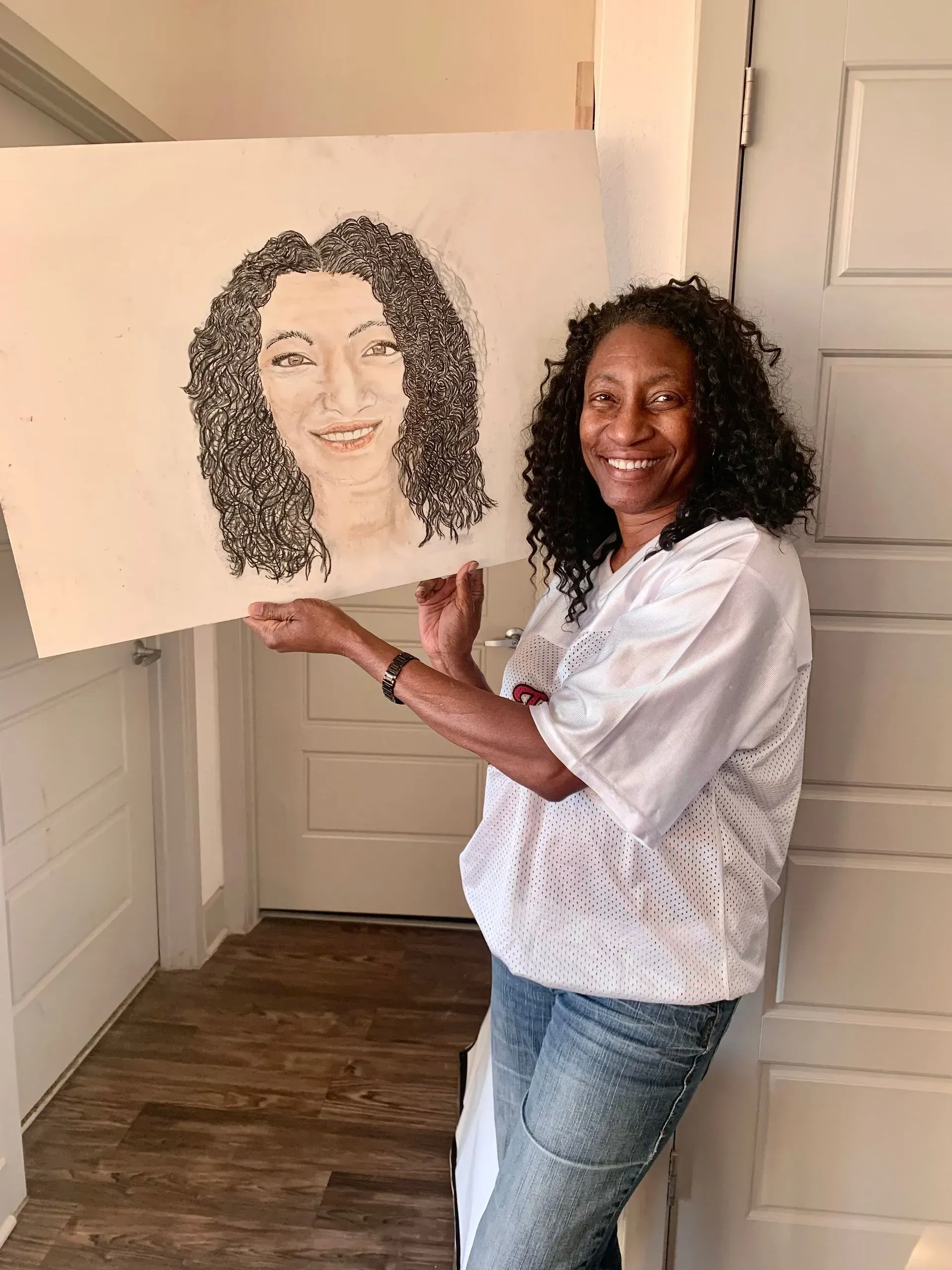 Woman holding a portrait she drew, smiling. She has curly hair and wears jeans and a white top. Indoors, in a doorway.