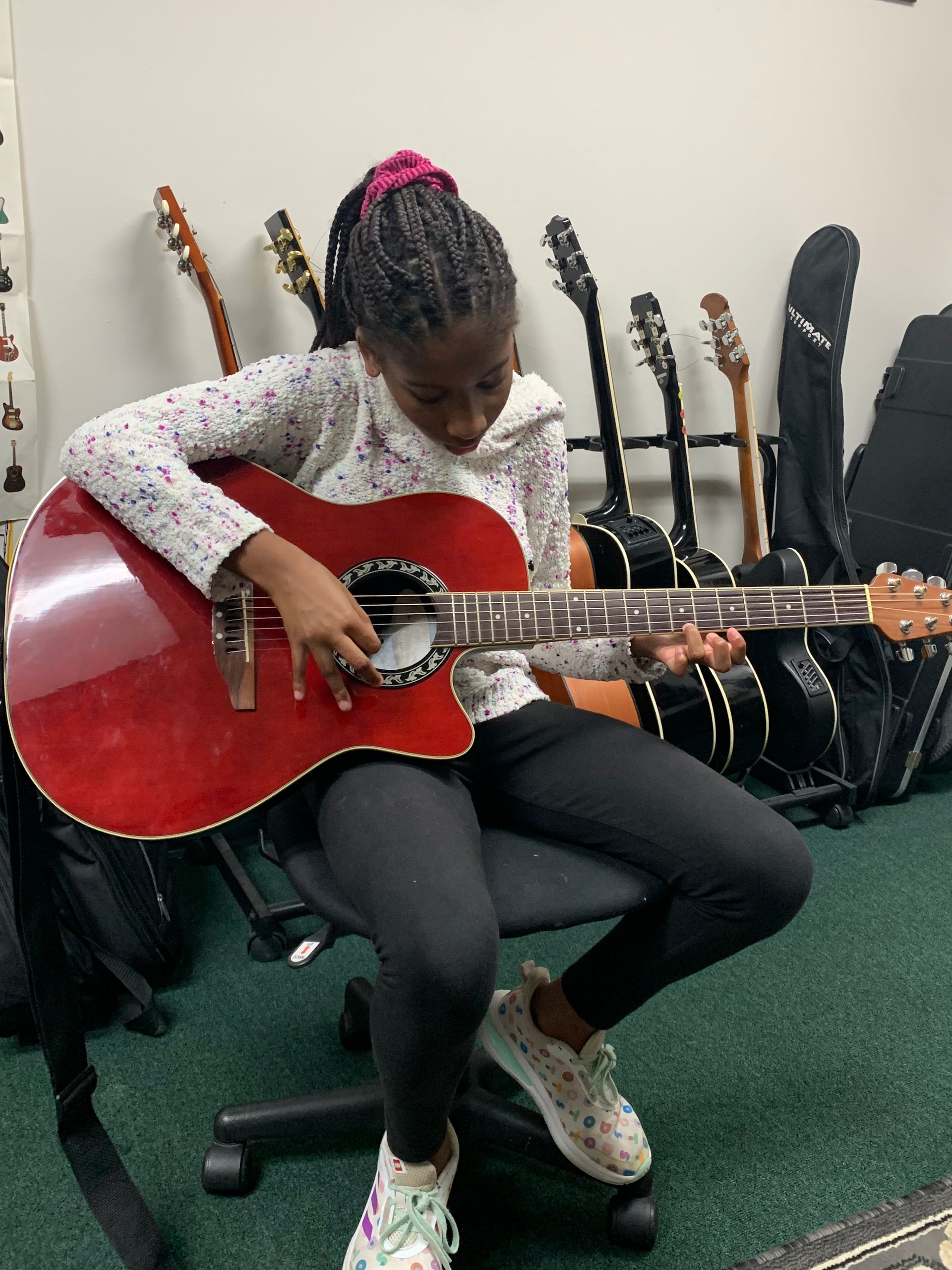 Girl playing a red acoustic guitar, seated. Background includes other guitars.