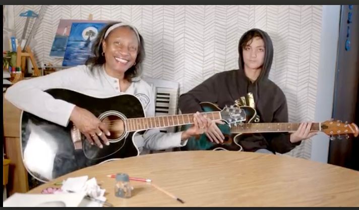 Woman and teen playing guitars, smiling, indoors, with painting and keyboard in background.