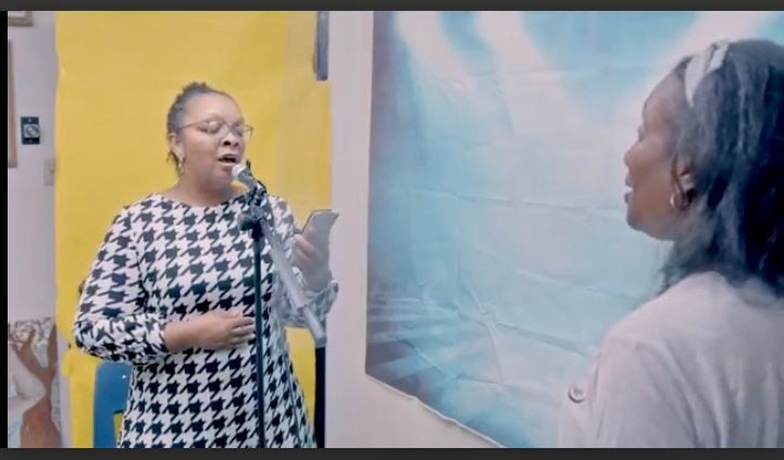 Woman in black and white patterned dress singing into a microphone in a studio; another woman watches.