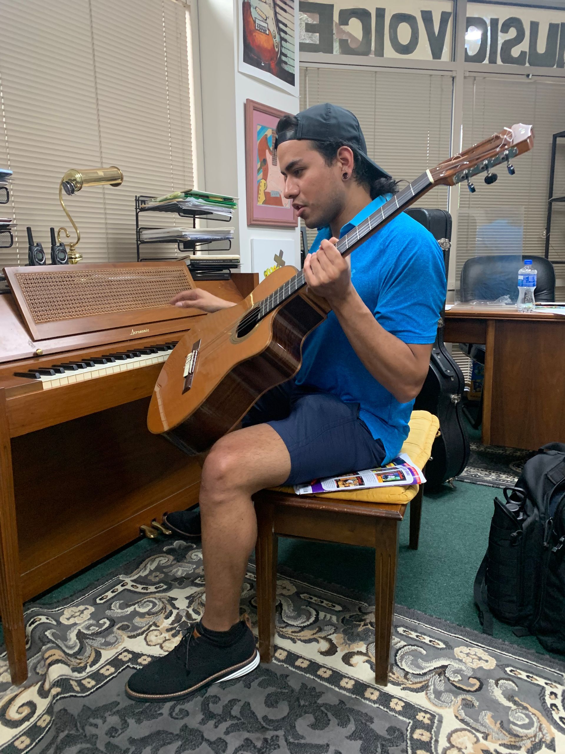 Man playing a 12-string guitar in a music studio with a piano. He wears a blue shirt, shorts, and a cap.