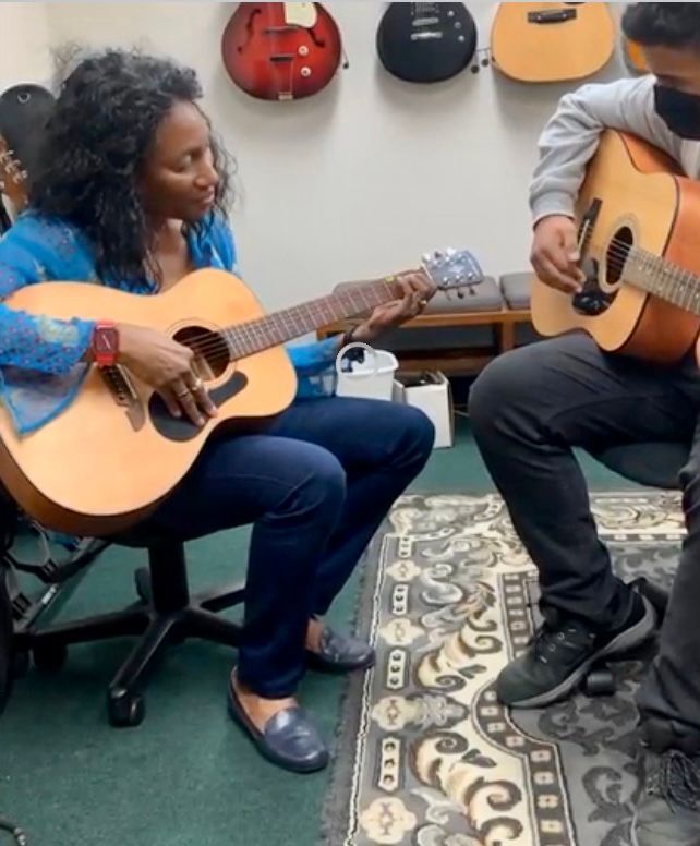 Woman and person playing acoustic guitars in a room with guitar displays.