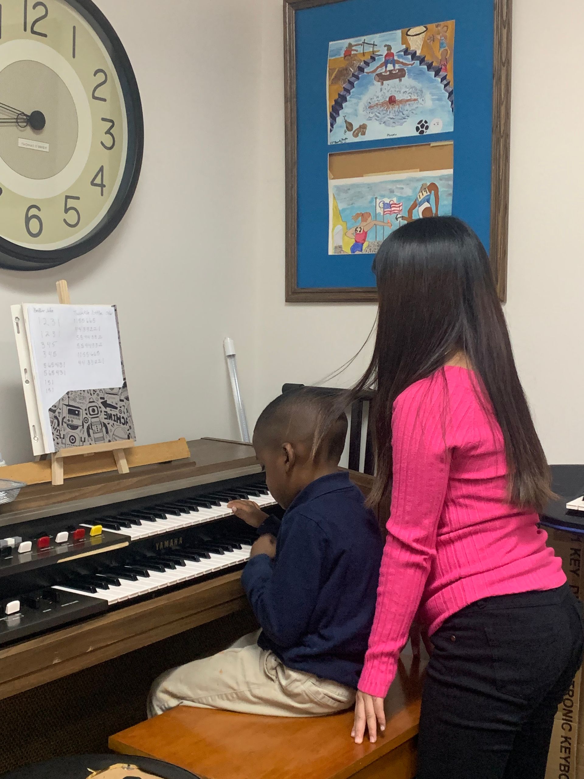 Woman guides child at an organ. Child plays keys, looking down. A clock and framed art are on the wall.