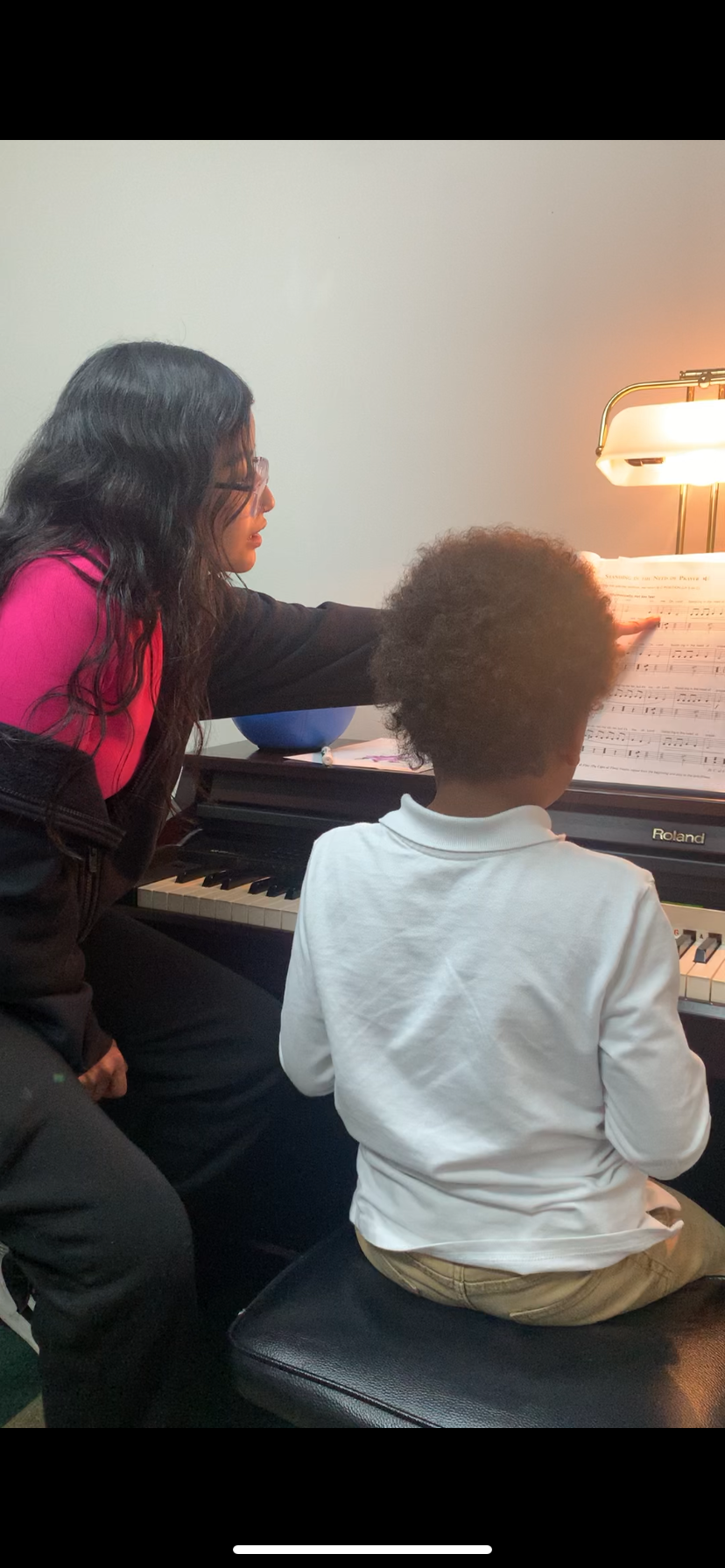 A music teacher guides a child at a piano, pointing to sheet music in a well-lit room.
