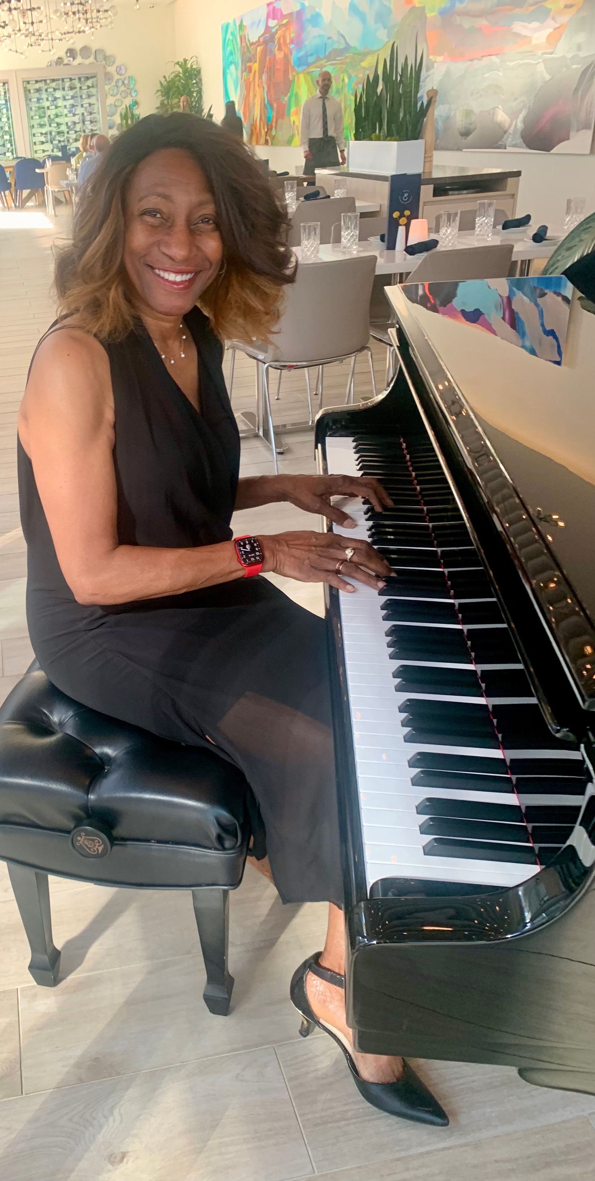 Woman playing a black piano, smiling in a bright room. She wears a black dress and heels.