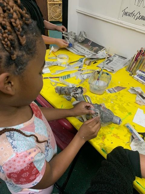 Girl with braided hair adds paper to a sculpture on a yellow table, others craft nearby.