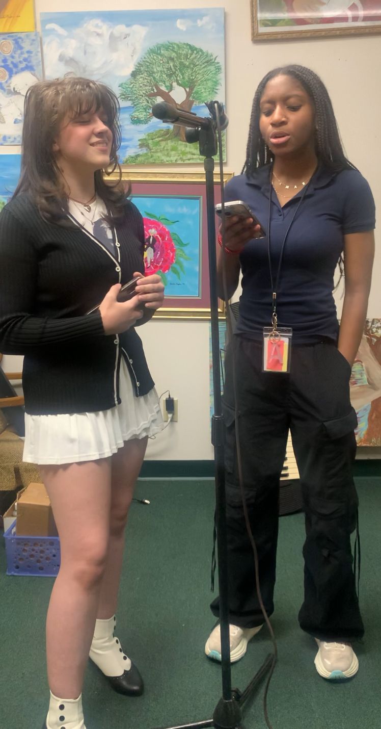 Two young women in a room with a microphone; one sings, the other holds a phone, possibly reading lyrics.