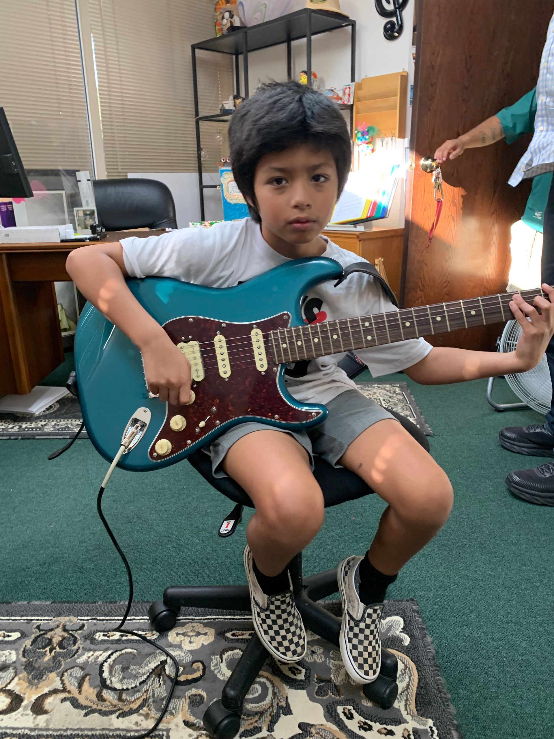 Boy seated, playing blue electric guitar. Wearing gray shorts, checkered shoes. Indoor setting.