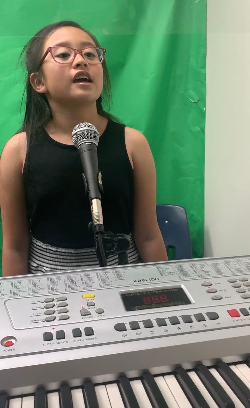 Girl singing into a microphone at a keyboard, wearing glasses and black top, green screen background.