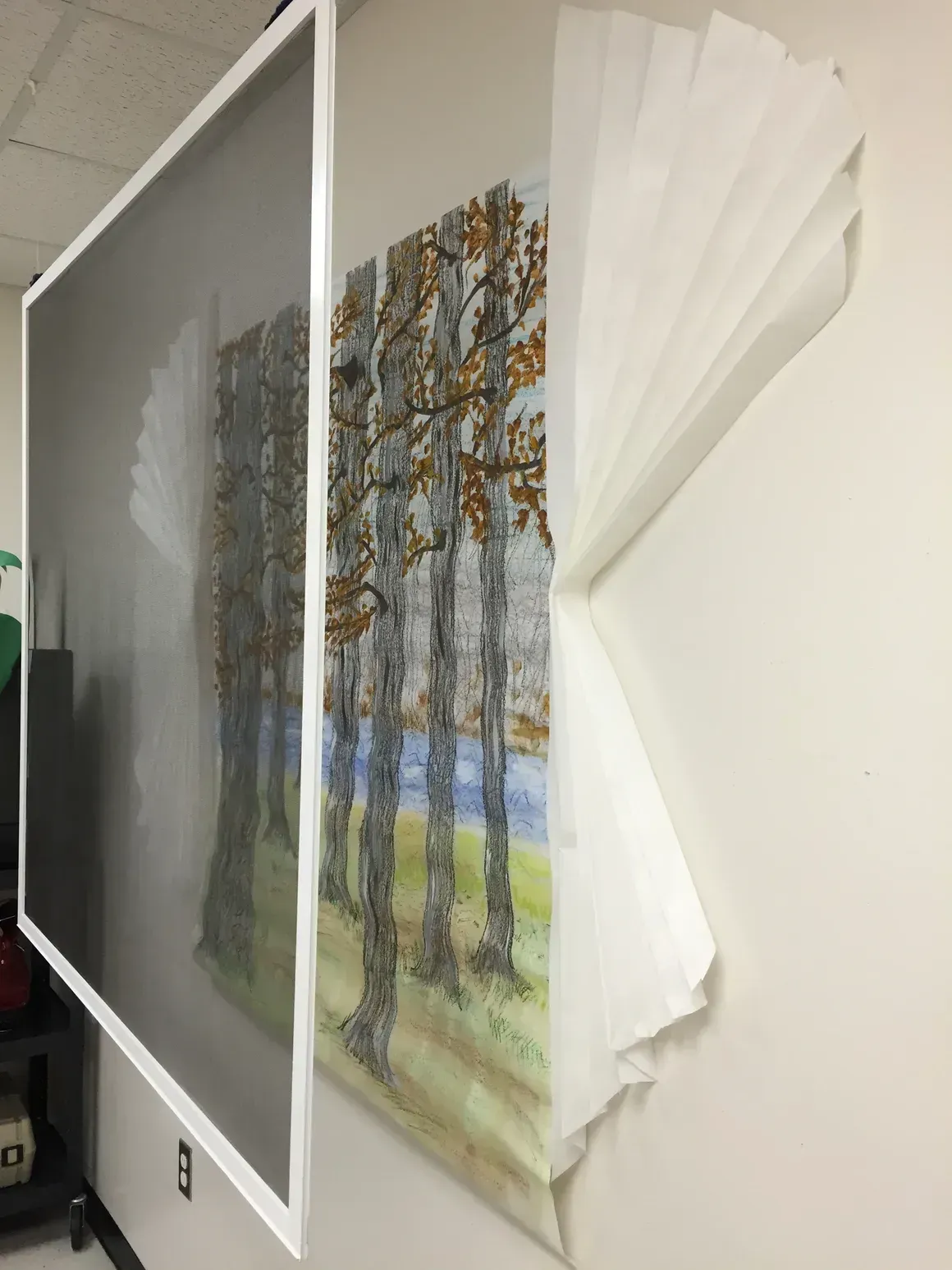 Artwork of a forest scene with layered paper and a screen overlay on a white wall.