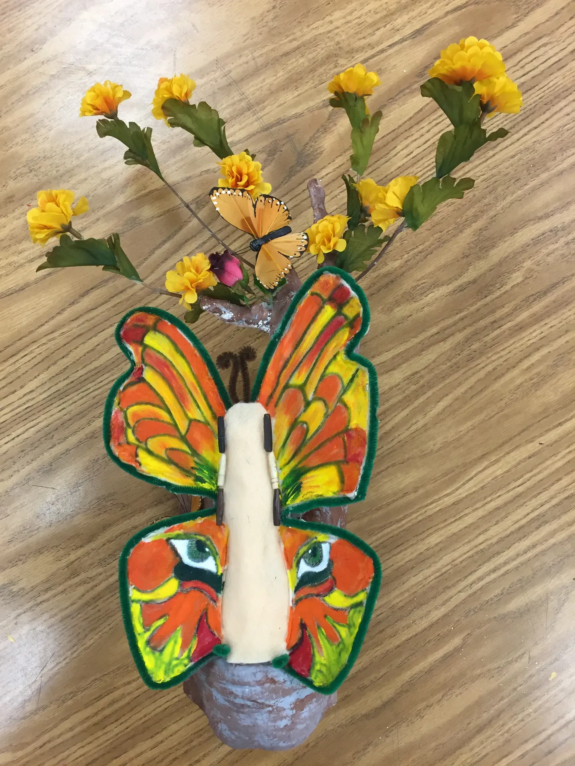 Butterfly-shaped vase with yellow flowers and a butterfly decoration on a wooden surface.