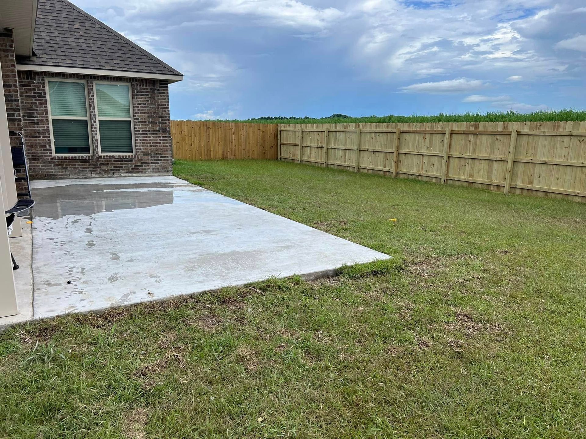 A backyard with a concrete patio, green grass, and a wooden fence under a cloudy sky.