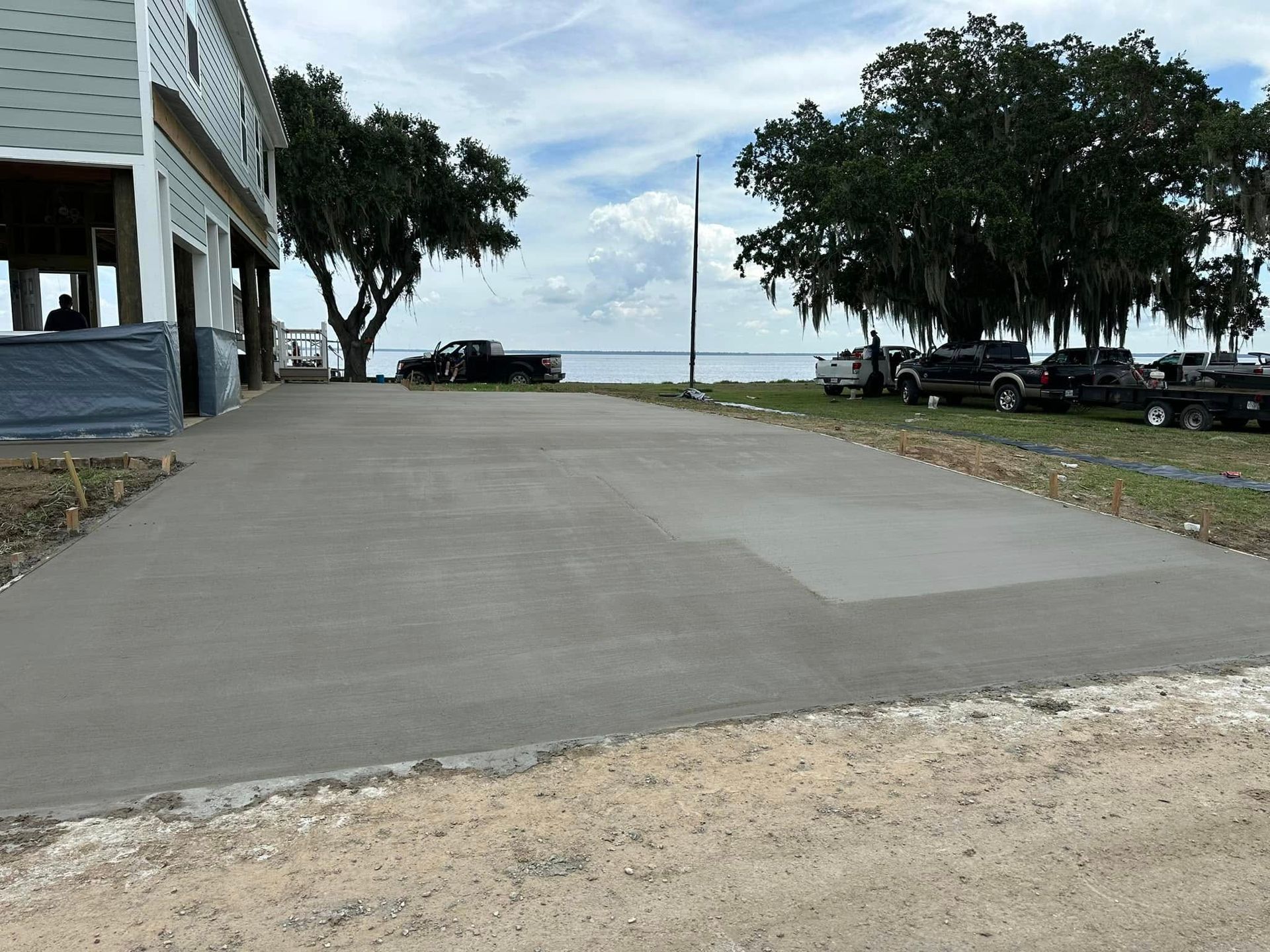 A newly poured concrete driveway leads to a waterfront, trees, and parked vehicles.