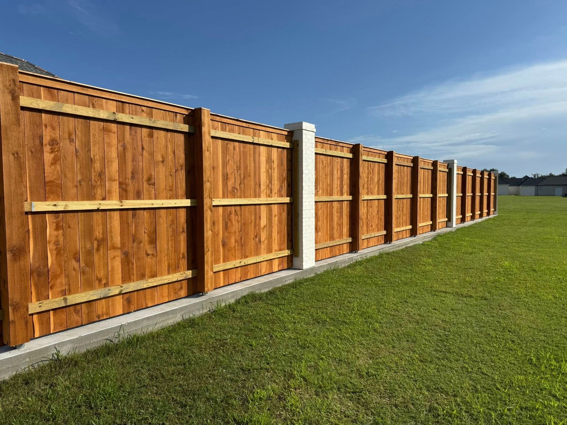 Wooden privacy fence with stained planks and white concrete posts in a grassy yard under a blue sky.