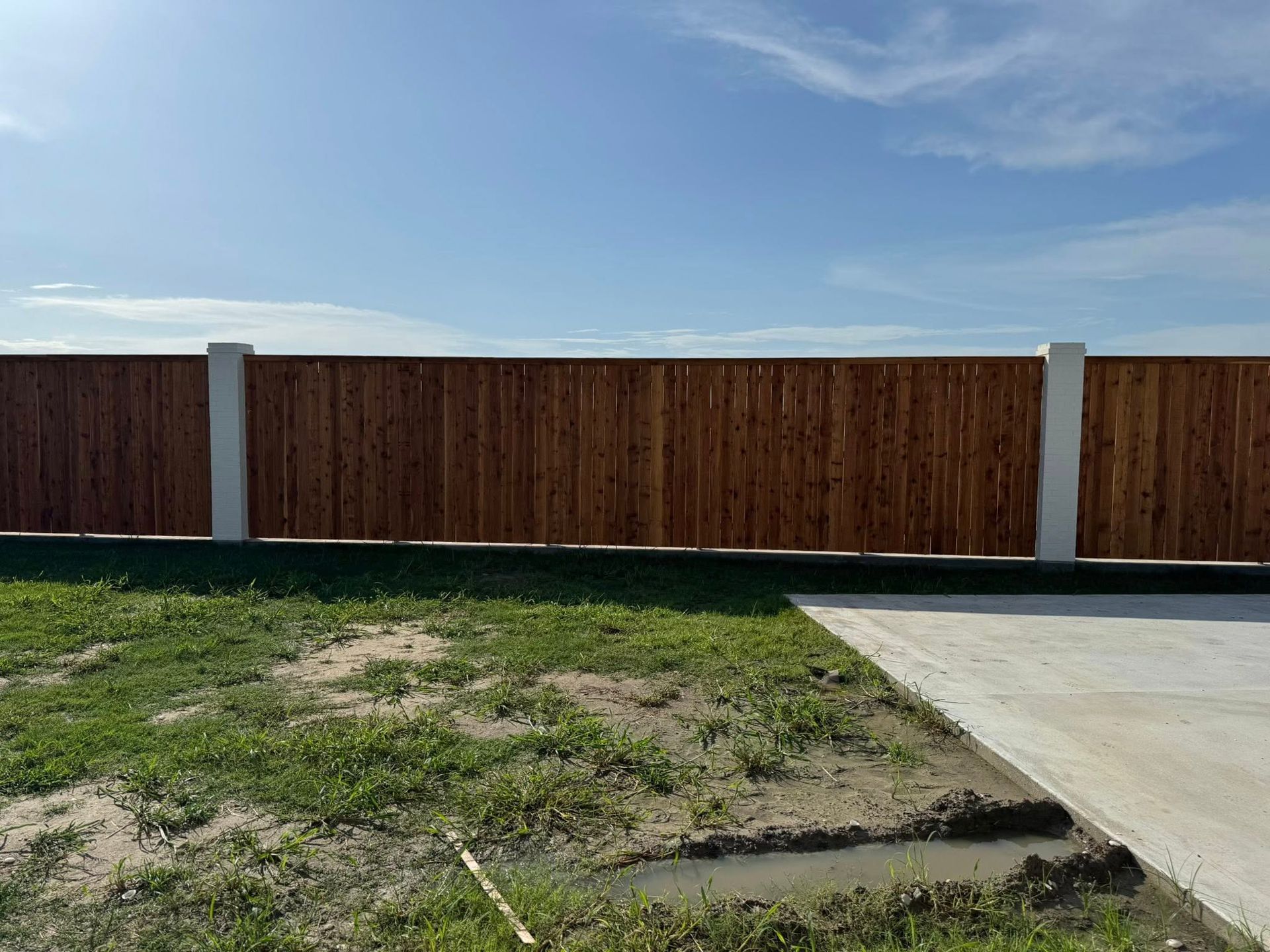 Wooden fence with white pillars, along a grassy lawn and concrete path, blue sky.