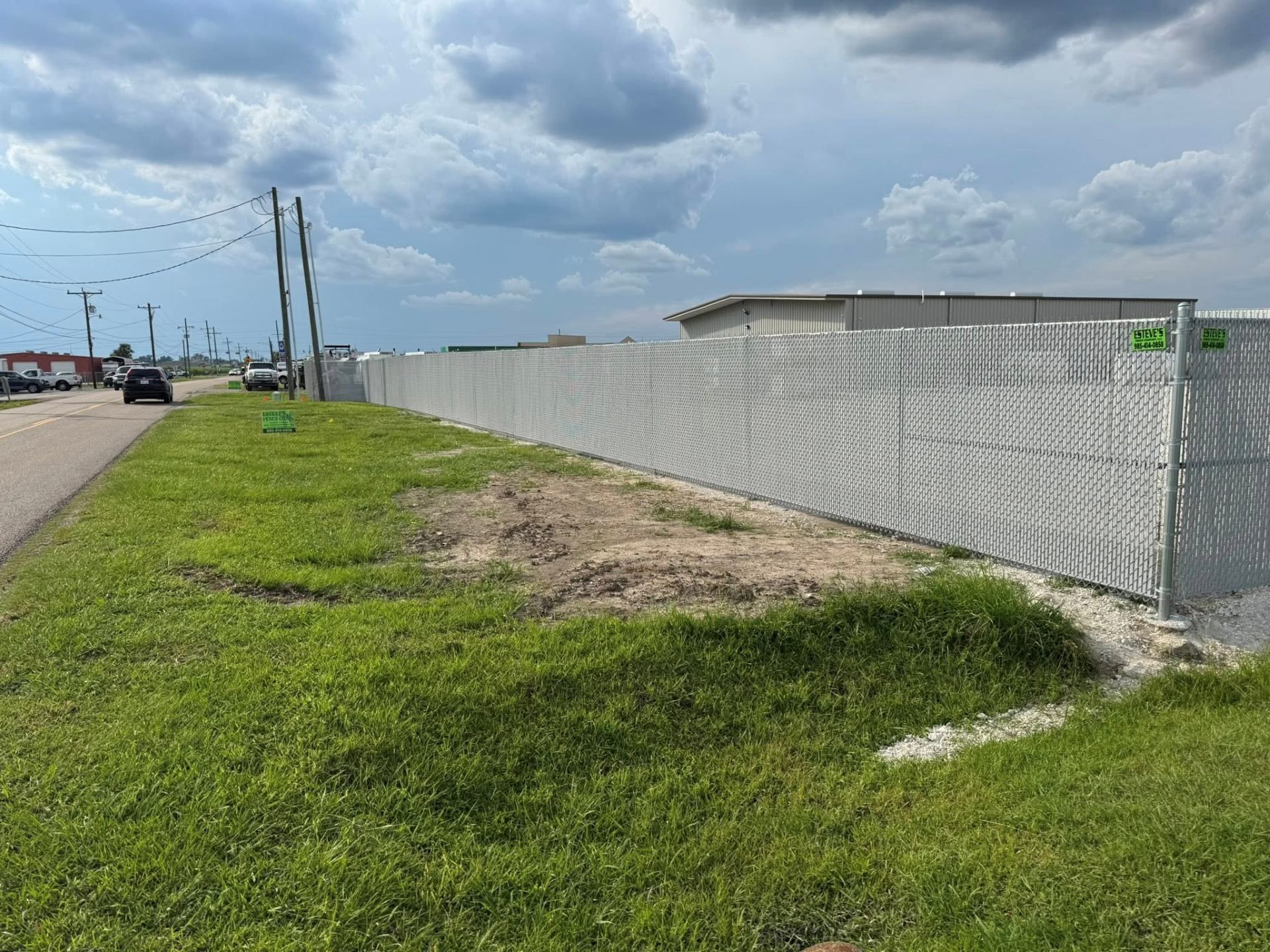 A long, silver chain-link fence along a grassy roadside with a cloudy sky overhead.