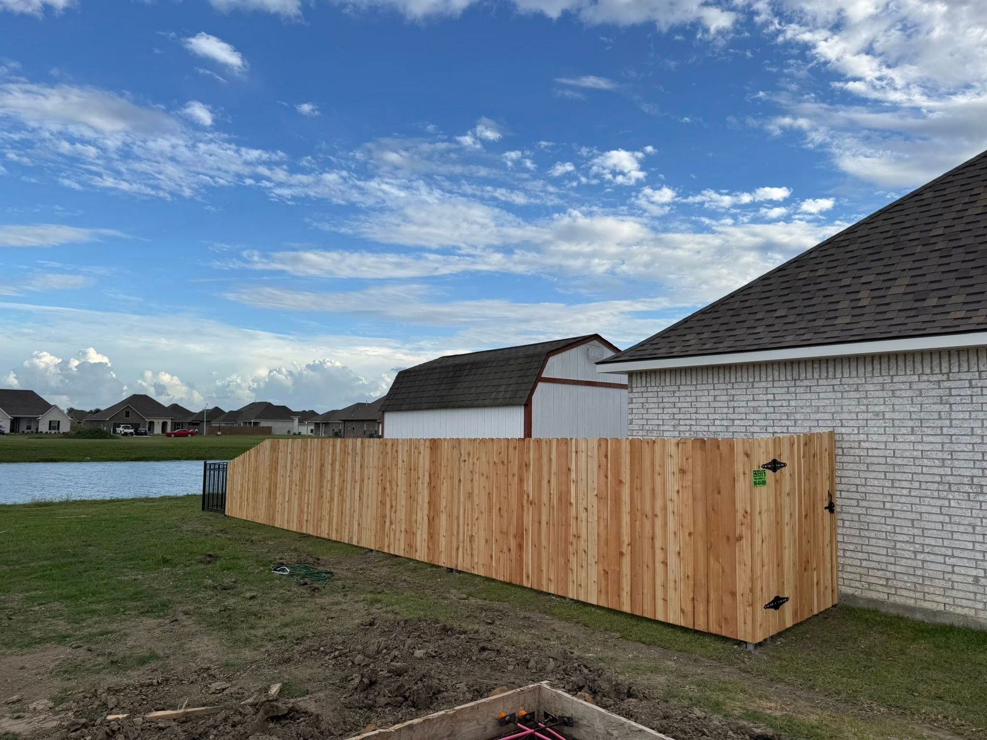 Wooden fence along a house and next to a grassy area, pond, and blue sky with clouds.